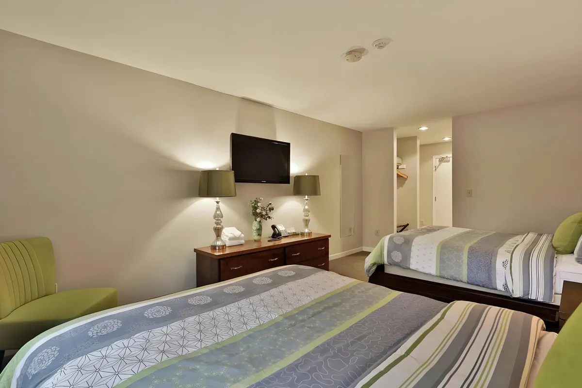 A comfortable bedroom featuring two beds with striped bedding, a wooden dresser, green-shaded lamps, and a wall-mounted television. The room feels spacious and is ideal for guests or kids, with ample storage and room for unwinding.