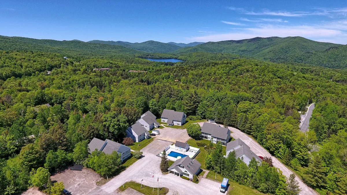 A sunlit outdoor swimming pool is framed by residential buildings and tall trees, with a dramatic mountain backdrop. The clear blue water invites swimmers, while the surrounding forest hints at nearby hiking trails and nature adventures.
