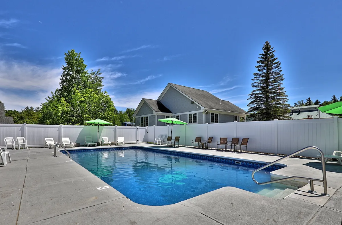 The swimming pool area features green umbrellas, lounge chairs, and is bordered by lush trees, evoking a tranquil and leisurely vibe. The inviting atmosphere suggests both relaxation and social gatherings by the water.