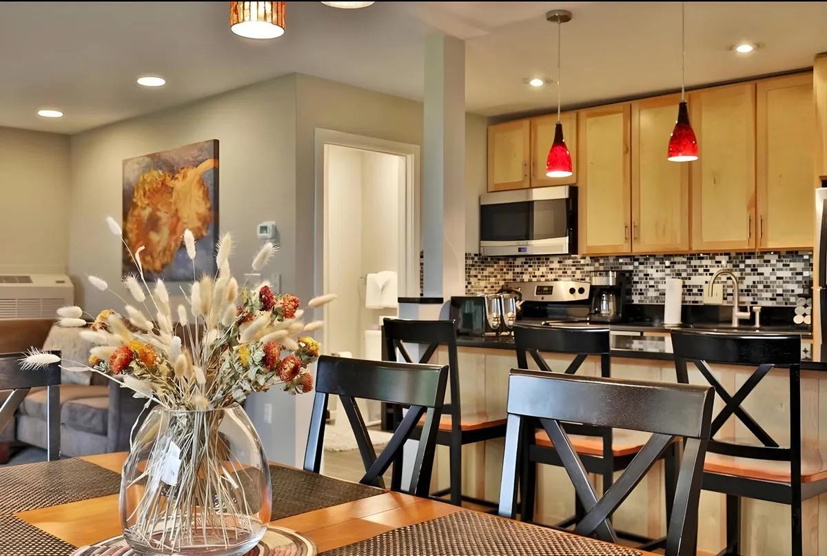 The image showcases a spacious and inviting dining area centered around a table with a vibrant floral arrangement, seamlessly connected to a modern kitchen with stainless steel appliances. Warm lighting and tasteful artwork set a cozy, welcoming mood, perfect for gatherings, shared meals, and relaxed mornings.