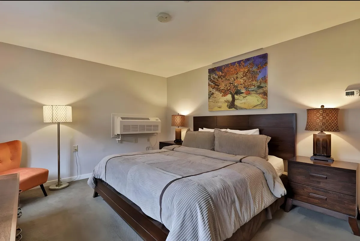 A serene bedroom with a king bed dressed in neutral, soft bedding, a colorful painting above the headboard, and an inviting accent chair positioned by the window. The room feels open, calm, and private—showcasing thoughtful design aimed at restful sleep and relaxation.