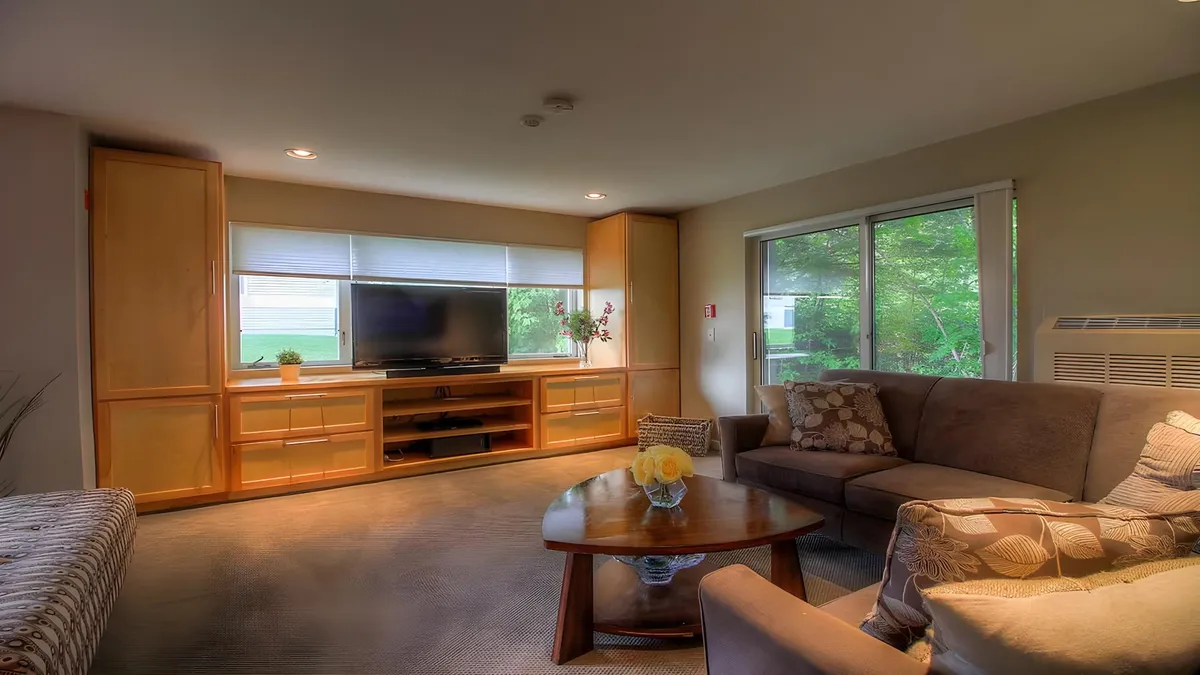 A cozy living room with plush seating arranged around a coffee table, a large smart TV set in a stylish unit, and natural light streaming in through sizable windows. The space exudes warmth and comfort, ideal for both relaxing and gathering with friends or family.