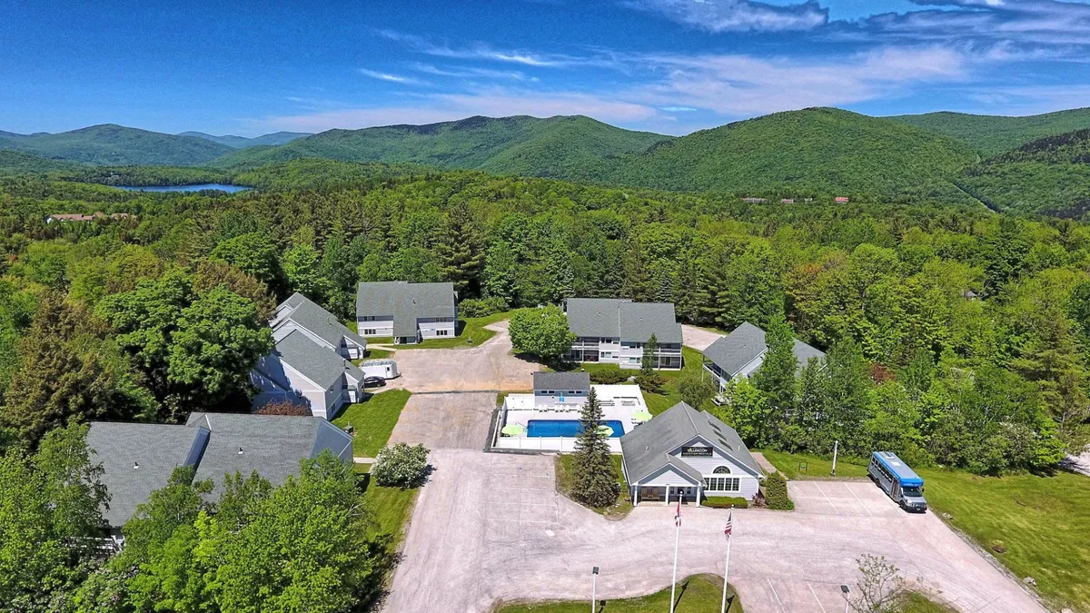 A sweeping exterior view of the swimming pool framed by residential buildings and landscaped gardens, all set against a dramatic mountain backdrop. The tranquil setting suggests both relaxation and the potential for group gatherings or family fun.