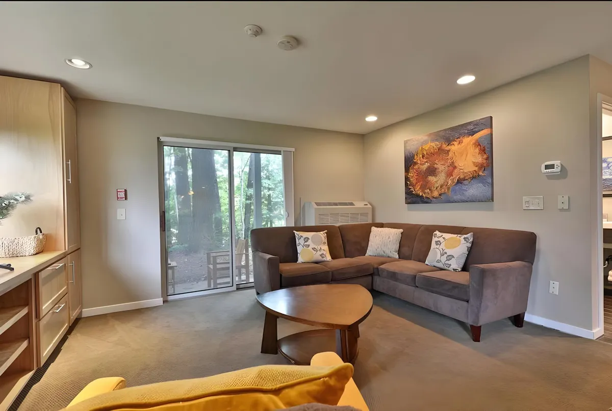This cozy living area features a sofa set, artwork on the walls, a coffee table, and a sliding glass door that opens to nature views. The natural light streaming in through the glass door draws attention to the seamless transition between indoor comfort and the beautiful outdoor environment.