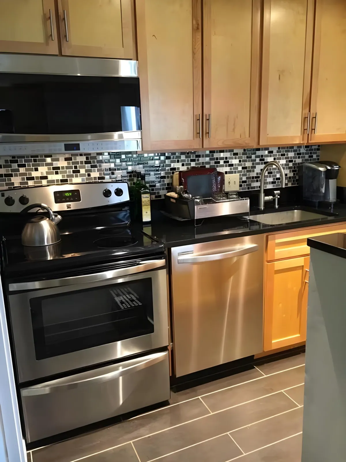 A modern kitchen with stainless steel appliances, black granite countertops, a colorful mosaic backsplash, and a kettle on the stove. The layout is practical and visually appealing, signaling a well-equipped culinary space.