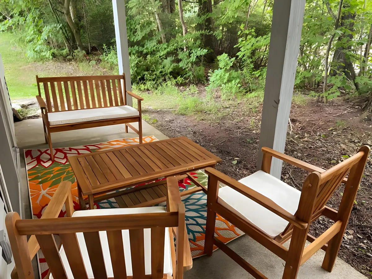 An outdoor seating area with wooden furniture arranged atop a colorful rug, surrounded by lush greenery. The scene suggests a tranquil spot for relaxing or socializing as the sun sets, seamlessly blending the comforts of home with the surrounding environment.