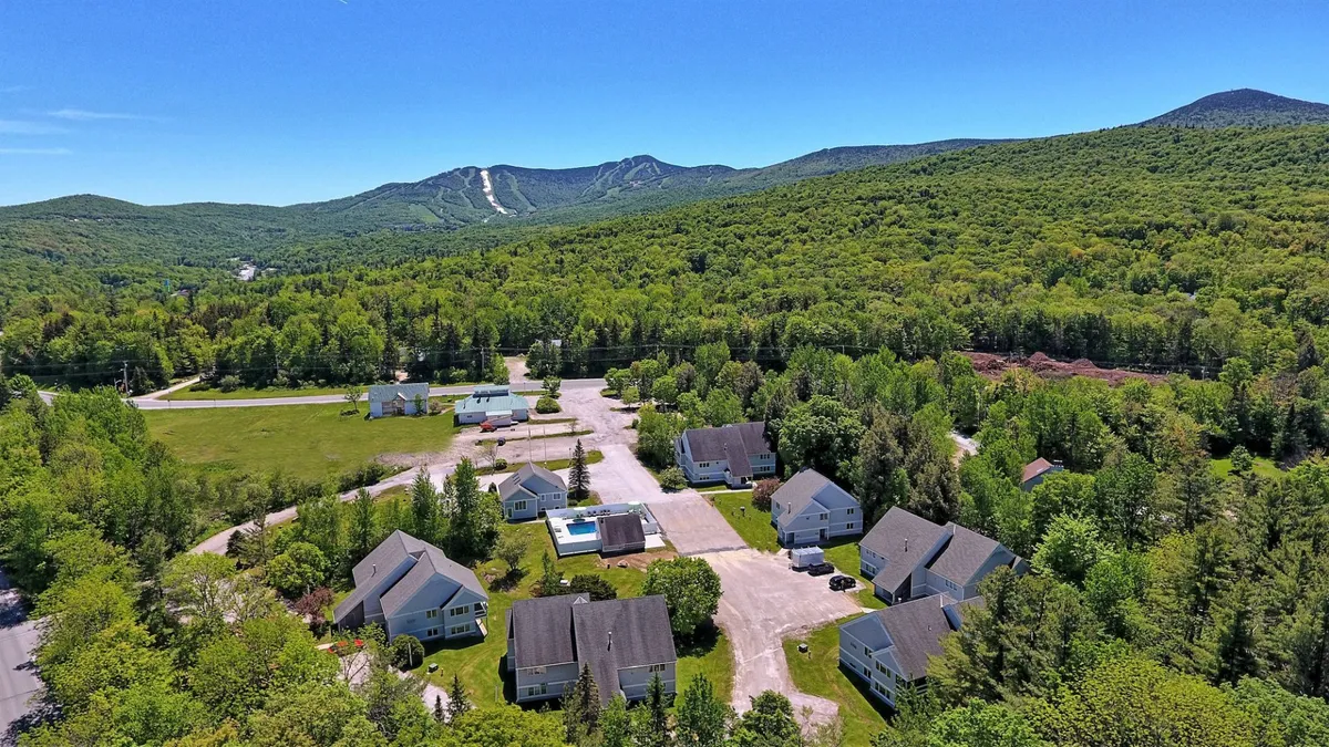 A wide exterior shot featuring a mountain range in the background, a cluster of houses nestled at the base, open green spaces, and a swimming pool. The landscape conveys a sense of proximity to the mountains and outdoor recreational opportunities, with the resort-like amenities anchoring the community to its natural surroundings.