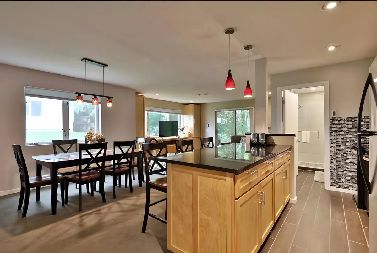 A spacious, open-plan kitchen featuring a central kitchen island, a dining table nearby, and decorative pendant lighting. Large windows fill the space with natural light, and the setup encourages cooking, dining, and socializing in one inviting area.